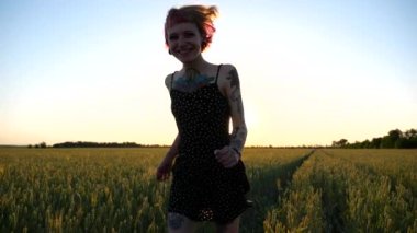 Güzel gün batımında buğday çayırında koşan dövmeli mutlu punk kadın. Pembe saçlı neşeli bayan hippi yeşil arpa tarlasında koşarken eğleniyor. Hippi kız yaz gününün tadını çıkarıyor..