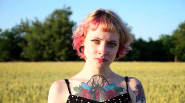 Arka planda bulanık buğday yeşili tarlası olan, kulak tünelleri olan mutsuz punk kızı portresi. Pembe saçlı ciddi hippi kadın gün batımında arpa çayırında kameraya bakıyor. Kapat..