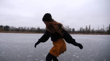 Kız bulutlu bir günde donmuş nehirde ya da gölde paten kayıyor. Doğada buz pateni pistinde kayan artistik paten giyen genç bir kadın. Soğuk kış havasında sporcu kadın eğitimi. Kış zamanı aktif bir eğlence. Yavaş çekim.