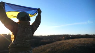 Üniformalı ve miğferli kadın asker tepede bayrak kaldırdı. Ukrayna askeri sıhhiyesi Ukrayna bayrağı sallıyor. Rus saldırısına karşı zafer. İstila direnci konsepti. Yavaş çekim.