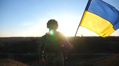 Soldier of ukrainian army stands at hill and holds waving flag of Ukraine. Man in military uniform and helmet lifted up flag against sunset. Victory against russian aggression. Invasion resistance.
