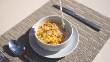 Detail view on stream of fresh milk pouring into bowl with dry crispy cornflakes. Close up of bowl with healthy breakfast and cutlery on table. Healthy food concept. Slow motion.