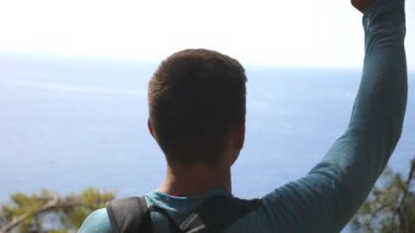 Sırt çantalı neşeli yürüyüşçü dağın yamacında zaferle el kaldırıyor. Erkek turist yolculuk boyunca aktif olarak dinleniyor ve özgürlüğün tadını çıkarıyor. Deniz manzaralı güzel bir manzara. Yavaş çekim