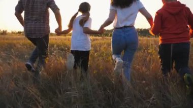 Mutlu, genç bir aile, birbirlerinin kollarını tutuyor ve çayırlarda koşuyor. Anne ve baba ve iki çocuk gün batımında çim tarlasında koşuyorlar. Çocuklarıyla birlikte hafta sonunun tadını çıkaran ebeveynler..