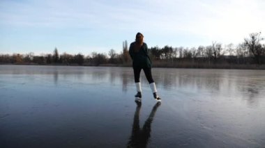 Gün batımında donmuş nehirde kayan genç bir kız. Artistik patenlerle buz pateni pistinde kayan kadın patenci. Soğuk havada gölde sporcu kadın eğitimi. Kış zamanı aktif bir eğlence. Yavaş çekim.