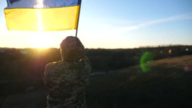Ukrayna ordusunun askeri tepede duruyor ve elinde Ukrayna bayrağı sallıyor. Askeri üniformalı tanınmayan adam gün batımına karşı bayrak kaldırdı. Rus saldırısına karşı zafer. İstila direnişi
