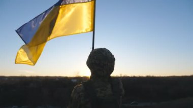 Askeri üniformalı genç adam Ukrayna bayrağını gün batımının arka planında sallıyor. Ukraynalı erkek asker ulusal bayrağı kaldırıyor. Rus saldırganlığına karşı zafer. Arka görünüm.