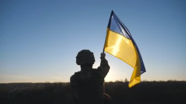 Askeri üniformalı erkek askerin Dolly çekimi Ukrayna bayrağını sallayarak tepenin zirvesinde duruyor. Ukraynalı ordu mensubu, günbatımının arka planında ulusal bayrağı kaldırdı. İstila direnişi kavramı.