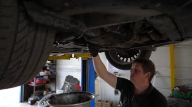 Profesyonel tamirci üniformalı. Garajdaki arabanın altındaki kirli yağı boşaltıyor. Genç tamirci atölyede kaldırma aracının altında çalışıyor. Otomobil bakım kavramı.