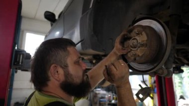 Yetişkin erkek tamirci atölyede kaldırma aracının altında çalışıyor. Garajda tornavida kullanan profesyonel sakallı otomobil tamircisi. Otomobil bakım kavramı.