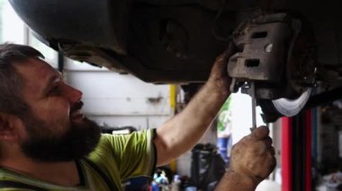 Üniformalı erkek oto tamircisi atölyede bir kaldırma aracının altında çalışıyor. Yetişkin profesyonel tamirci serviste ya da garajda araba tamir ediyor. Düzenli otomobil bakımı kavramı. Yavaş çekim.