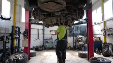 Üniformalı bir tamirci atölyedeki bir arabanın süspansiyonunu inceliyor. Garajda kaldırma aracının altında çalışan profesyonel sakallı oto tamircisi. Otomobil hizmeti kavramı. Yavaş çekim.