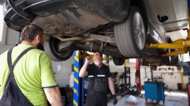 Garajda teknik inceleme ve araç kontrolü yapan profesyonel tamirciler. Steteskopla dinleyen erkek oto tamircisi atölyedeki kaldırma otomobilinin altında makine parçası arızası.