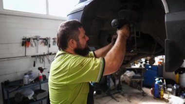 Atölyedeki özel elektrikli aletle erkek tamirci cıvataları sıkılaştırıyor. Profesyonel oto tamircisi garajdaki kaldırma aracının altında çalışır. Yetişkin tamirci kapalı alanda ulaşımı tamir ediyor.