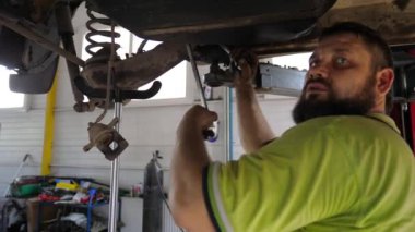 Üniformalı araba tamircisi atölyedeki ingiliz anahtarı aletini kullanarak aracın tamirini yapıyor. Yetişkin erkek tamirci garajdaki kaldırma aracının altında çalışıyor. Otomobil bakımı kavramı.