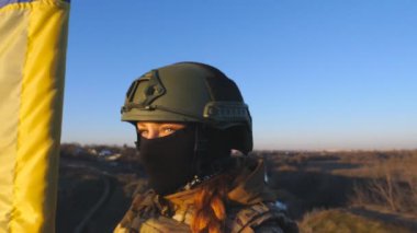 Ukraynalı asker kız günbatımının arka planında ulusal bayrağı kaldırıyor. Askeri üniformalı genç bayan asker tepenin zirvesinde Ukrayna bayrağı sallıyor. Savaşın zaferi. Dolly vurdu..
