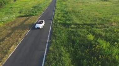 Boş kırsal yolda giderken çekilen beyaz üstü açık araba görüntüsü. Cabriolet 'te gezen tanınmayan dört genç kadın. Yaz günü kırsal yolda otomobil sürerken. Yavaş çekim.