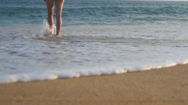 Güneşli bir günde sığ sulara girerken okyanus dalgalarıyla yıkanan kadın ayakları. Yüzmek için denize yürüyen genç kadın bacakları. Kız kıyı şeridinde vakit geçiriyor. Yaz tatili veya tatil kavramı.
