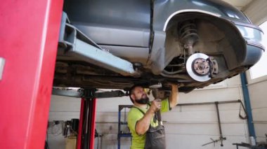 Yetişkin erkek tamirci garajdaki bir kaldırma aracının altında çalışıyor. Üniformalı profesyonel sakallı tamirci atölyede çekiç kullanan arabanın tamirini yapıyor. Otomobil bakımı kavramı.