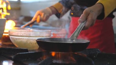 Tanımlanamayan şef iki tava tutuyor ve modern otel mutfağında flambe tarzı yemekler pişiriyor. Erkek aşçı fırında sebzeleri karıştırıp kızartıyor. Yemek pişirme kavramı. Ağır çekim kenar görünümü