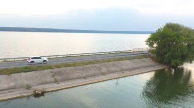 Göl kenarındaki yolda uçan arabanın görüntüsü. Beyaz SUV yaz günü baraj yolunda gidiyor. Otomatik olarak nehir ya da gölet köprüsünden geçiyor. Manzaralı bir yer. Seyahat ve tatil kavramı. Üst görünüm