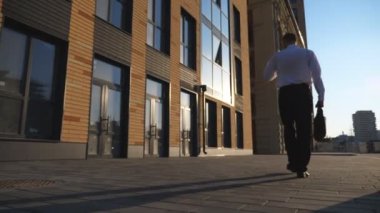 Evrak çantalı genç işadamı cep telefonuyla kaldırımda işe gidiyor. Başarılı bir girişimci resmi giyimde şehir caddesindeki ofis binasının yakınında akıllı telefon kullanıyor. Ağır çekim..