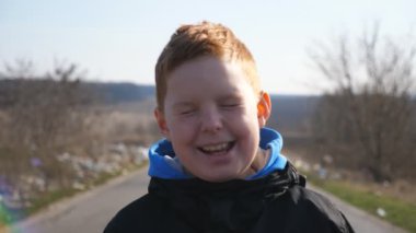 Gülümseyen, ceketli kızıl saçlı çocuğun portresi dışarıdaki kameraya bakıyor. Çilli küçük komik çocuk kırsalda yolda duruyor ve gülüyor. Arka plan bulanık. Yavaşça kapat..
