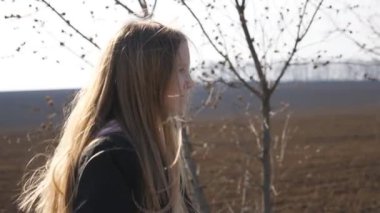 Little smiling girl with backpack goes along a driveway near ploughed meadow. Small kid moves on way to school at spring day. Happy female child with long blonde hair walks on road near plowed field.