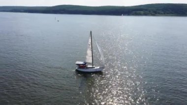 Açık mavi gökyüzü altında sakin bir göl boyunca ilerleyen birkaç yelkenlinin hava görüntüsü. Yatlar gölde yüzüyor ve arka planda güzel doğa manzarası var. Özgürlük ve sükunet kavramı. Yavaş çekim