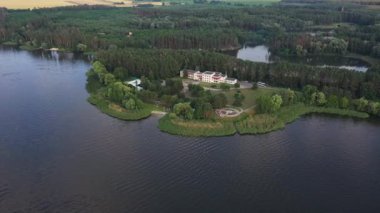 Gölle çevrili güzel bir yarımadanın havadan görünüşü. Etrafı yemyeşil ağaçlar ve yeşil çimlerle çevrili büyük bir malikane. Yaz gününde güzel manzaranın üzerinde uçmak. Yavaş çekim.
