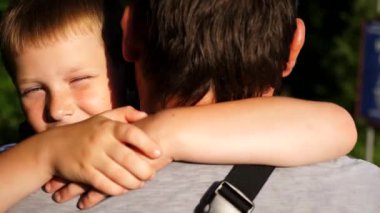 Küçük oğlunun elini tutan ve kırsalda yürüyen genç bir baba. Küçük çocuk kameraya bakıyor ve babasına sarılıyor. Güneşin parlak ışıklarıyla aydınlatan sevimli gülümseyen çocuğun yüzü.