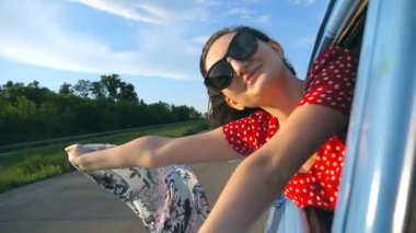 Güneş gözlüklü, elinde eşarpıyla pencereden sarkan ve keyifli bir seyahat yapan genç bir kız. Çekici bir kadın gülümser ve güneşli bir günde seyahatten hoşlanır. Seyahat konsepti. Yavaş çekim. Yakın çekim..