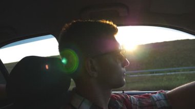 Profil arka plan güneş patlaması ile araba güneş gözlüklü genç adam. Yakışıklı olan günbatımında otomatik sürüş. Onun araç içindeki beyaz oğlan. Salon yan görünümden. Ağır çekim yakın çekim.