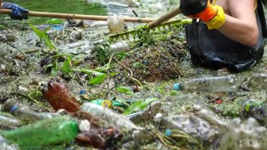 Koruyucu eldivenli kadın çevrecilerin elleri enkaz nehirlerini temizliyor. Kirli gölde plastik şişe toplayan kauçuk elbiseli tanınmayan gönüllüler. Çevre kirliliği koruması.