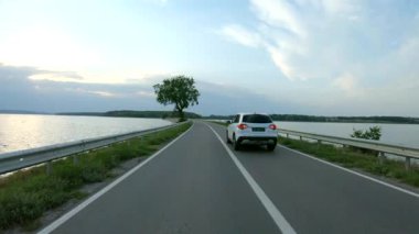 İnsansız hava aracı baraj yolundan geçen beyaz arabayı takip ediyor. SUV nehir ya da göl kenarındaki güzel kırsal yolda gidiyor. Yolculuk ya da doğa yolculuğu. Tatil aile seyahati kavramı. Havadan çekim Ağır çekim.