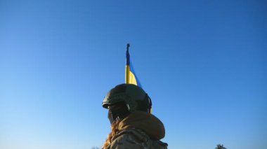 Ukrayna bayrağıyla tepenin zirvesinde duran Ukrayna ordusunun kadın ve erkek askeri. Kamuflaj üniformalı askeri çift, güneşli mavi gökyüzüne bakarak el ele tutuşuyorlar. Dolly vurdu.