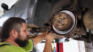 Üniformalı profesyonel sakallı tamirci atölyede çekiç kullanan arabanın tamirini yapıyor. Yetişkin erkek tamirci garajdaki bir kaldırma aracının altında çalışıyor. Otomobil bakımı kavramı.