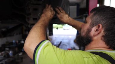 Üniformalı erkek oto tamircisi atölyede bir kaldırma aracının altında çalışıyor. Yetişkin profesyonel tamirci serviste ya da garajda araba tamir ediyor. Düzenli otomobil bakımı kavramı. Yavaş çekim.