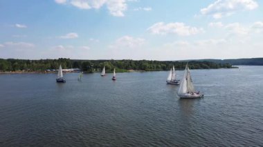 Açık mavi gökyüzü altında sakin bir göl boyunca ilerleyen birkaç yelkenlinin hava görüntüsü. Yatlar gölde yüzüyor ve arka planda güzel doğa manzarası var. Özgürlük ve sükunet kavramı. Yavaş çekim