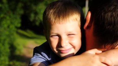 Küçük oğlunun elini tutan ve kırsalda yürüyen genç bir baba. Küçük çocuk kameraya bakıyor ve babasına sarılıyor. Güneşin parlak ışıklarıyla aydınlatan sevimli gülümseyen çocuğun yüzü.