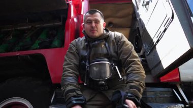 Portrait of happy male firefighter in uniform against background of a fire truck. Young fireguard looking into camera and smiling near a big red car. Concept of saving lives and heroic profession.