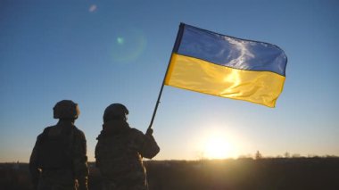 Ukrayna ordusunun genç askerleri ulusal bayrağıyla tepenin zirvesinde dikiliyorlar. Kamuflaj üniformalı kadın ve adam Ukrayna bayrağını kırsal kesimde kaldırarak gün batımına bakıyorlar. Savaşta zafer.
