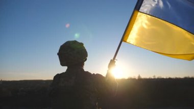 Ukraynalı bir asker tepede durup gün batımına karşı mavi-sarı pankart sallıyor. Askeri üniformalı erkek asker, Rus saldırısına karşı zafer sembolü olarak Ukrayna bayrağını kaldırıyor..