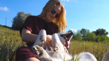 Sarı saçlı güzel bir kadın çimenlerde oturup Sibirya köpeğini okşuyor. Güneş gözlüklü genç kız, güneşli yaz gününde evcil hayvanıyla Meadow 'da vakit geçiriyor. Kapat..