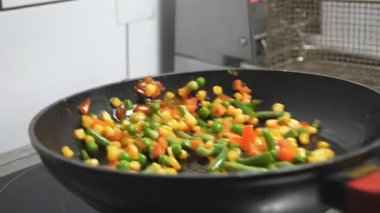 Aşçının erkek kolu tava tutuyor ve mutfak restoranında karışık sebzeleri atıyor. Aşçı kızarmış tavada mısır, bezelye, kuşkonmaz, biber ve havuç sote ediyor. Yemek pişirme kavramı.