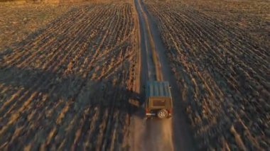 Tarlalar arasında boş kırsal yolda giden çiftçi cipinin hava görüntüsü. Alacakaranlıkta tozlu yolda giden off road aracı. Yaz günü kırsal kesimde araba sürerken. Üst görünüm.