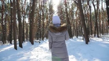 Back view of young woman walking in sunny winter forest. Unrecognizable girl enjoying stroll through snowy wood. Carefree lady going and admiring beautiful winter landscapes. Winter concept. Slow mo.