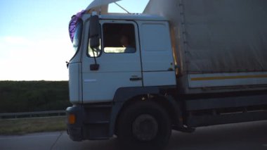 Kamyon arka planda gün batımına kadar kırsalda ilerliyor. Kamyon asfaltta gidiyor. Şoför yola dikkatlice bakıyor. Otoyolda giden bir kamyon. Yavaş çekim kenar görünümü.