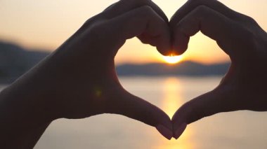 Genç kadın ellerinin silueti, güzel gün batımıyla deniz zemininde kalbi şekillendiriyor. Yaz tatili kavramı. Yavaşça kapat..