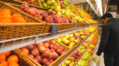 Genç adam renkli meyvelerle raf boyunca yürüyor ve elma seçiyor. Marketten ürün seçen bir erkek alıcı. Süpermarkette çeşitli ürünler. Alışveriş konsepti. Yavaş çekim.